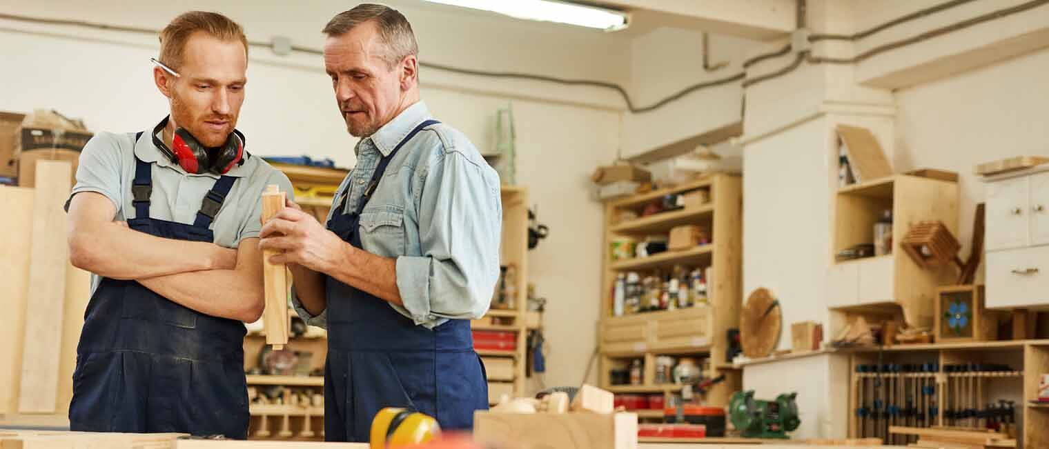 Unternehmensnachfolge Ein junger und ein älterer Handwerker stehen in einer Werkstatt und besprechen sich