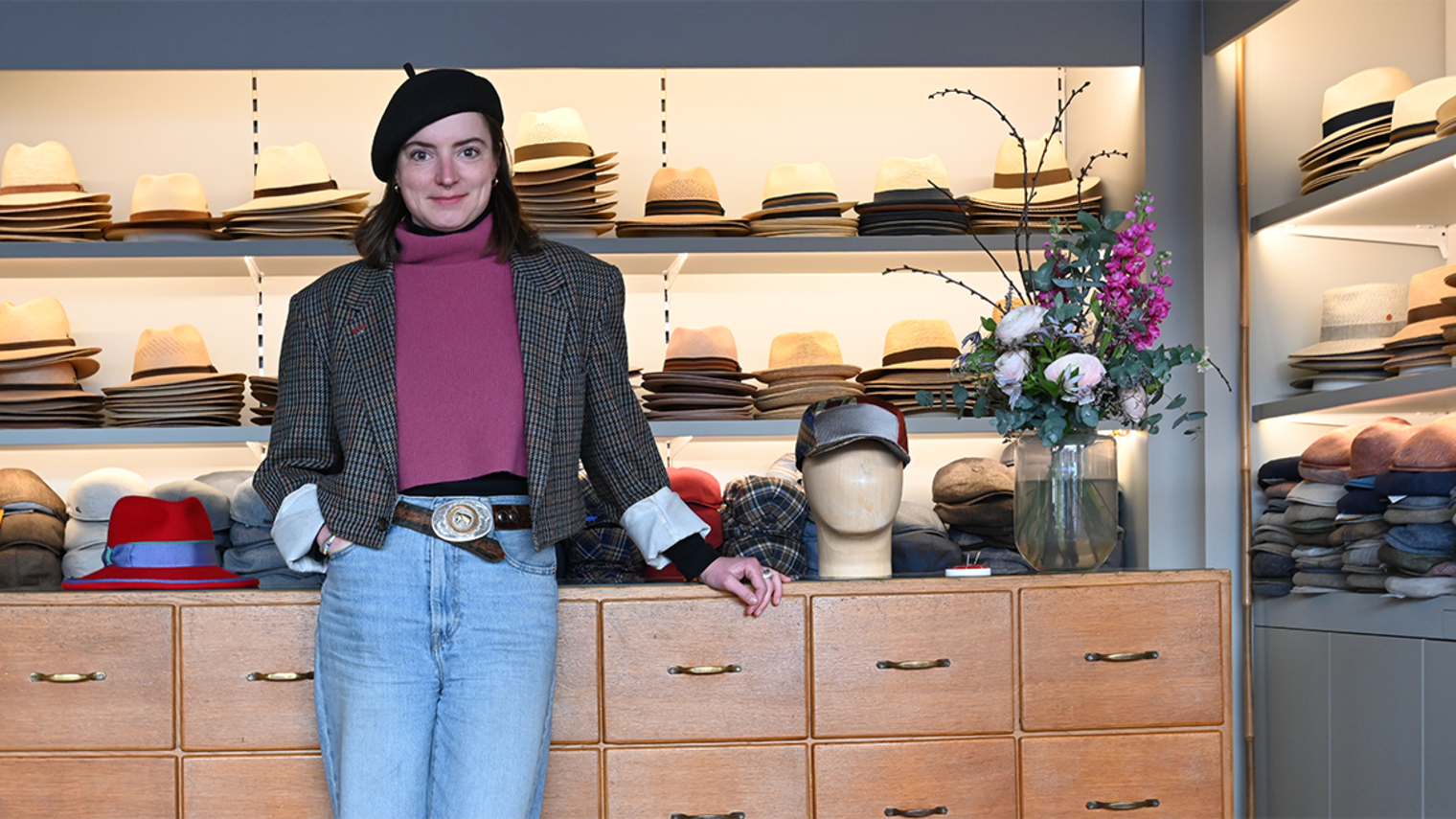 Modistin Laura Zieger in ihrem Geschäft in Würzburg vor einer Kommode, auf der Hüte liegen; im Hintergrund Regale mit Hüten.