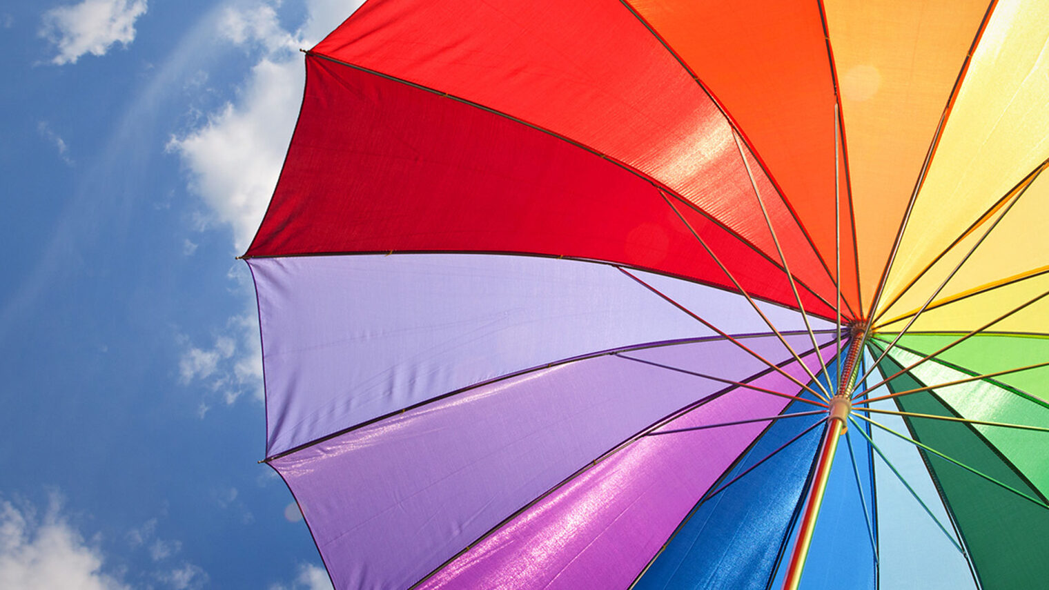 Bunter Sonnenschirm in Regenbogenfarben vor blauem Himmel mit Wolken