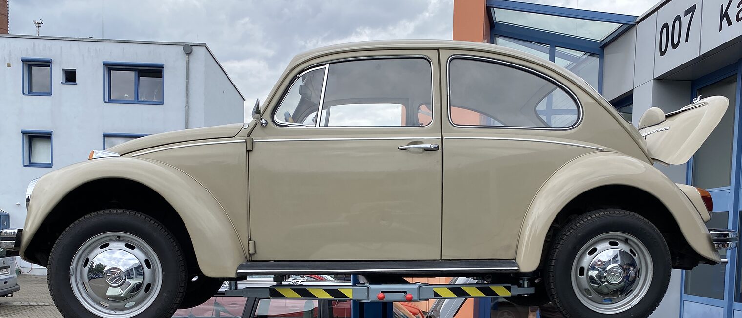 Beiger VW Käfer steht auf Hebebühne vor Werkstatt, darunter grauer Volvo, zwei Personen arbeiten am geöffneten Motorraum.