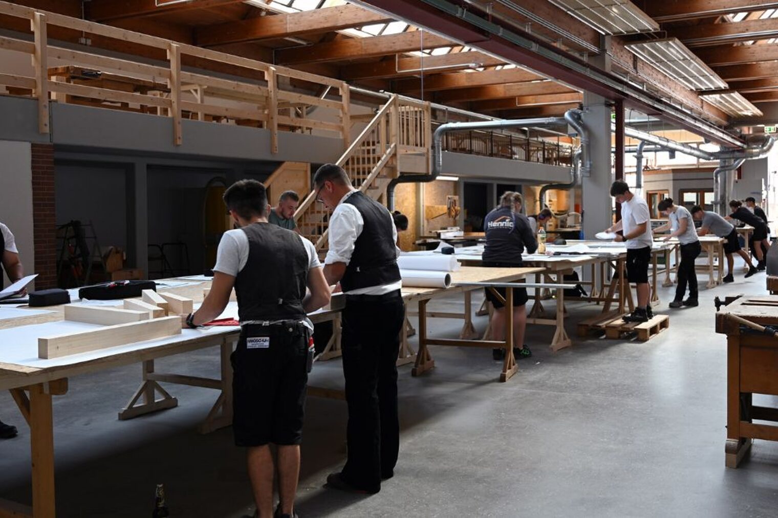 Zimmerer-Meisterschüler in Arbeitskleidung am Arbeiten an Tischen Große Halle mit mehreren Arbeitstischen, an denen Zimmerer-Meisterschüler in Arbeitskleidung und Kluft arbeiten
