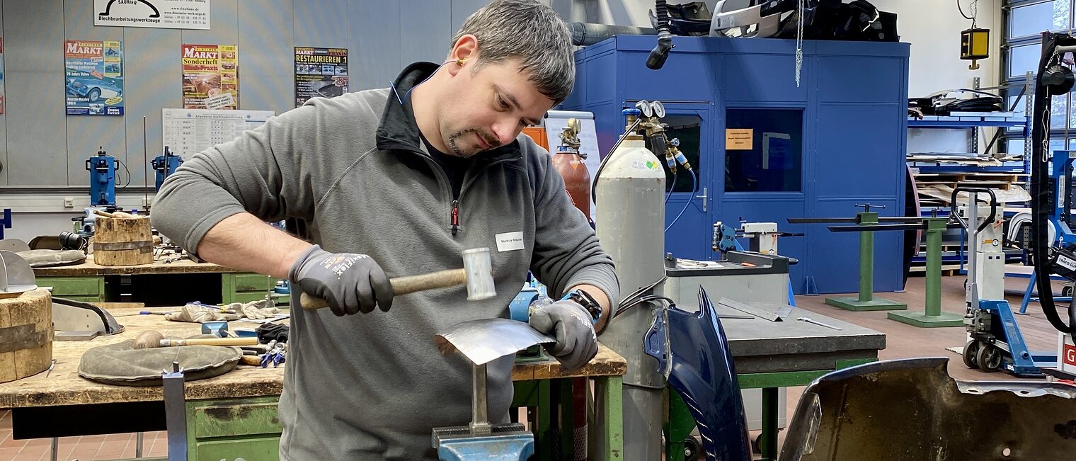 Ein Mann bearbeitet ein eingespanntes Metallblech mit Hammer, trägt Handschuhe, im Hintergrund Werkzeuge und Gasflaschen.