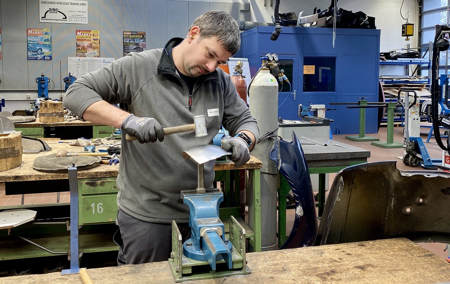 Ein Mann bearbeitet ein eingespanntes Metallblech mit Hammer, trägt Handschuhe, im Hintergrund Werkzeuge und Gasflaschen.