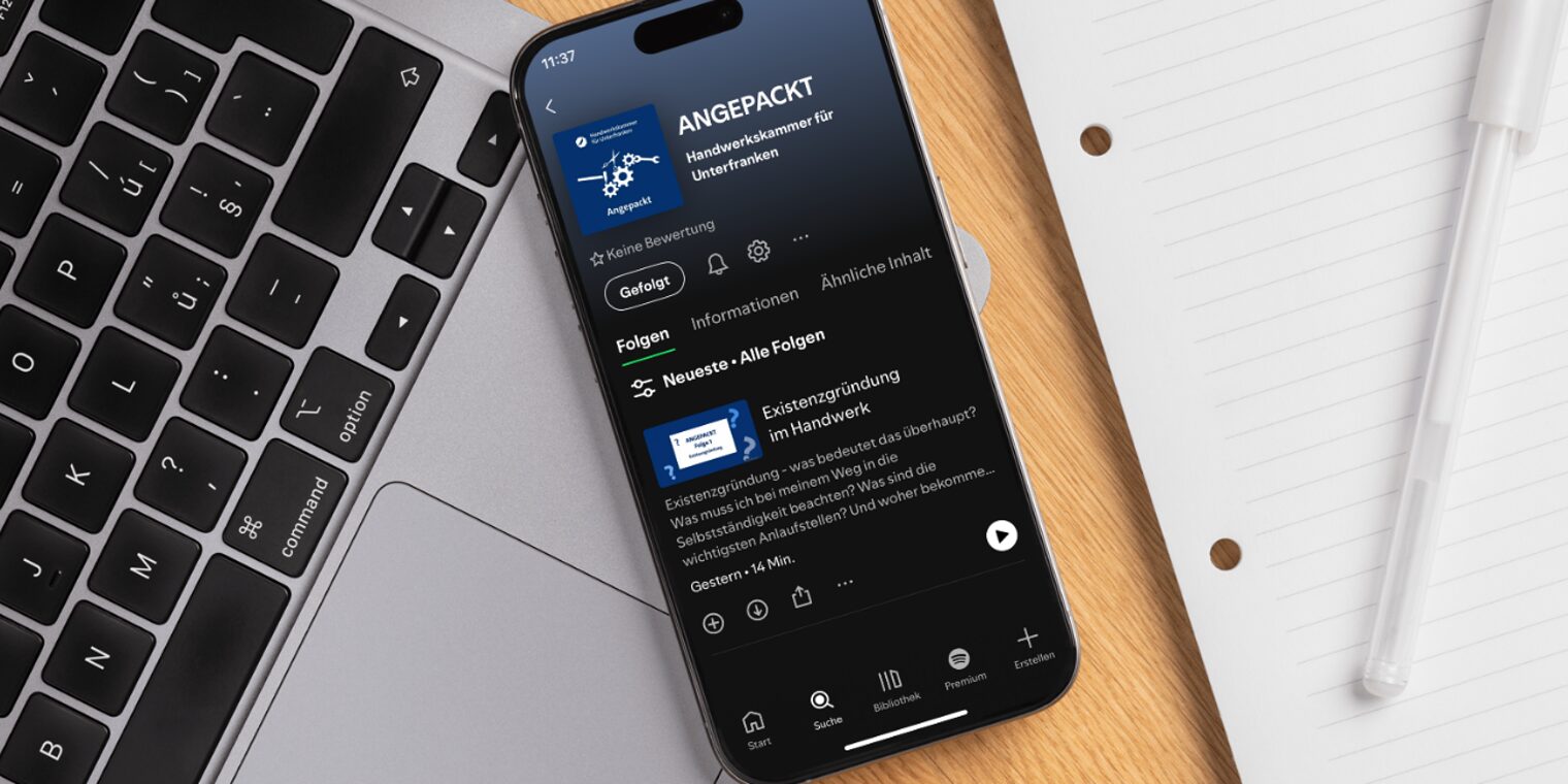 Smartphone mit Bildschirm, auf dem der Podcast "Angepackt" in Spotify geöffnet zu sehen ist, auf einem Schreibtisch.