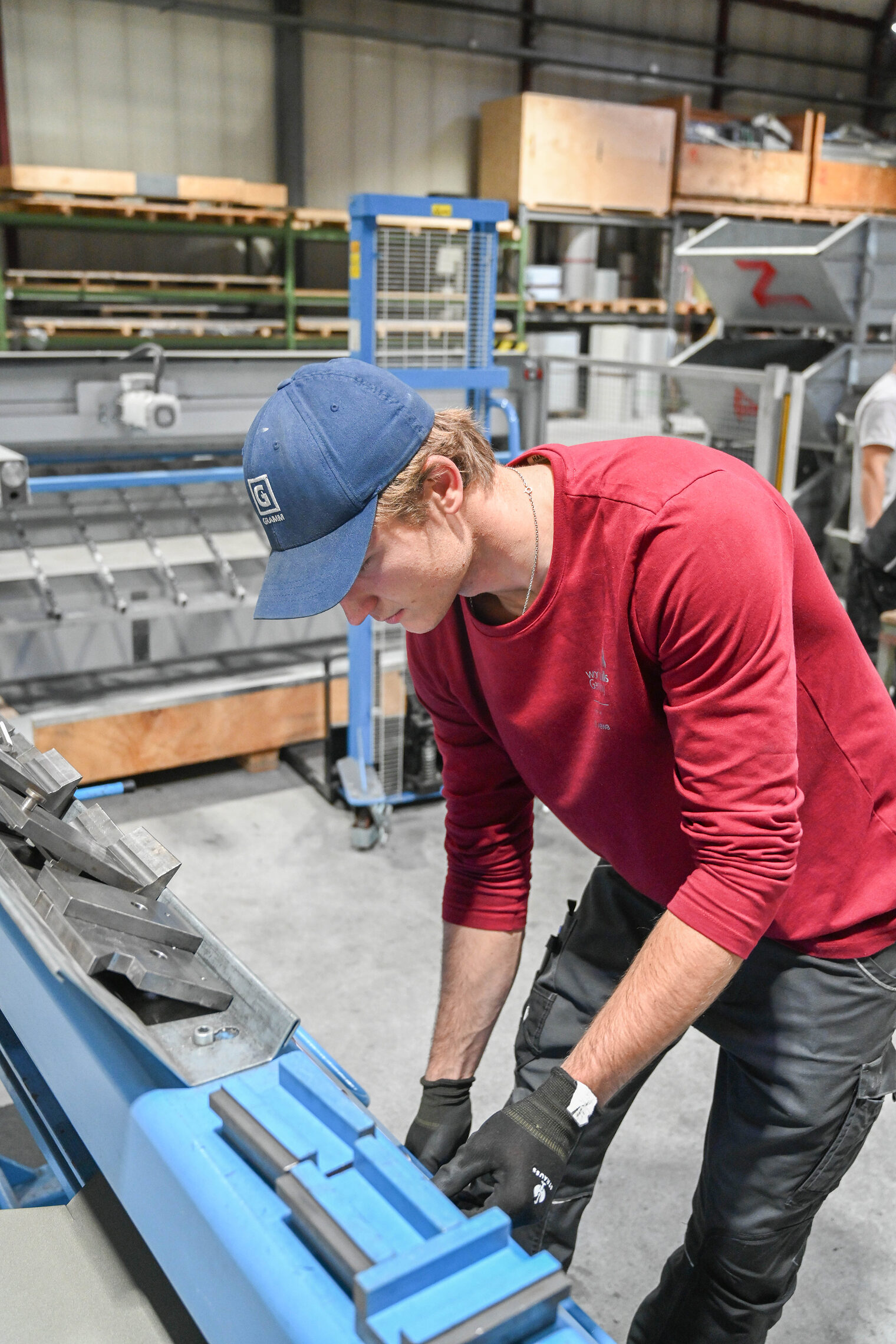 Junger Mann in rotem Longsleeve arbeitet mit Arbeitshandschuhen an einer Maschine zum Blechbiegen. 