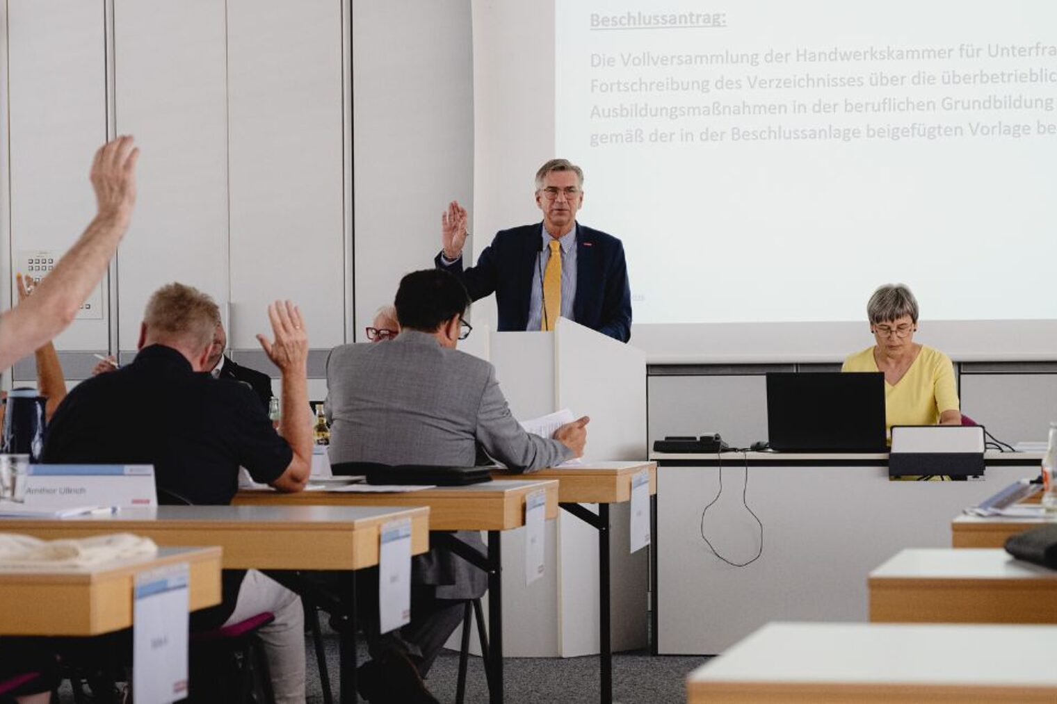 Mehrere Personen sitzen in einem Meeting-Raum und heben die Hände zur Abstimmung über einen Antrag