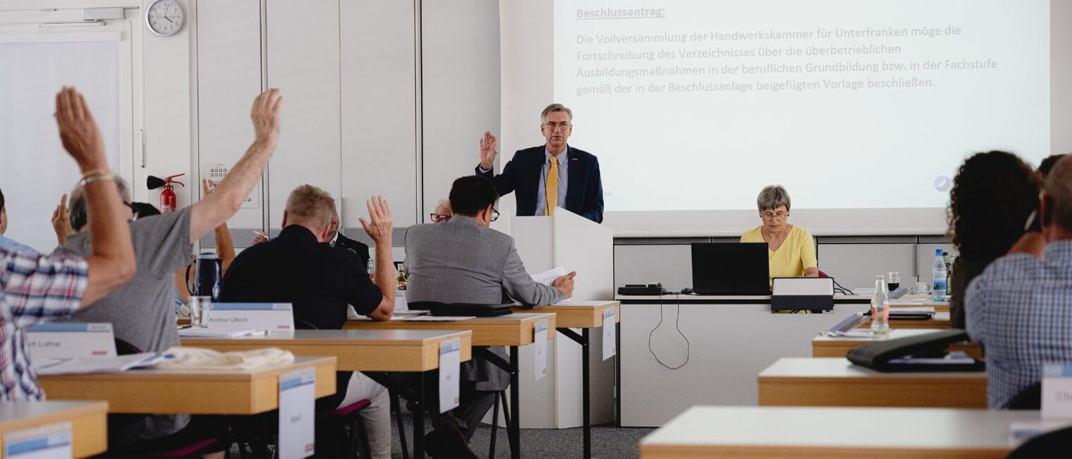Mehrere Personen sitzen in einem Meeting-Raum und heben die Hände zur Abstimmung über einen Antrag